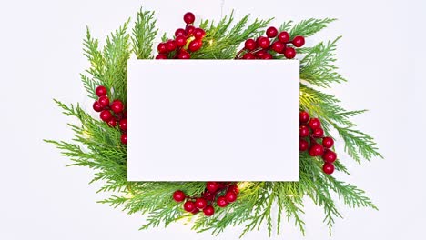 natural fir twigs with holly berries and blinking lights under white paper card note on white theme. stop motion