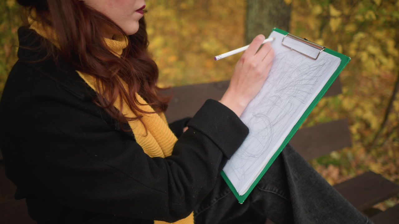Close up of young lady in sunglasses, yellow beret, and yellow muffler deeply focused on her drawing, sketching lines all over her canvas in peaceful outdoor autumn setting