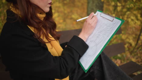 close up of young lady in sunglasses, yellow beret, and yellow muffler deeply focused on her drawing, sketching lines all over her canvas in peaceful outdoor autumn setting