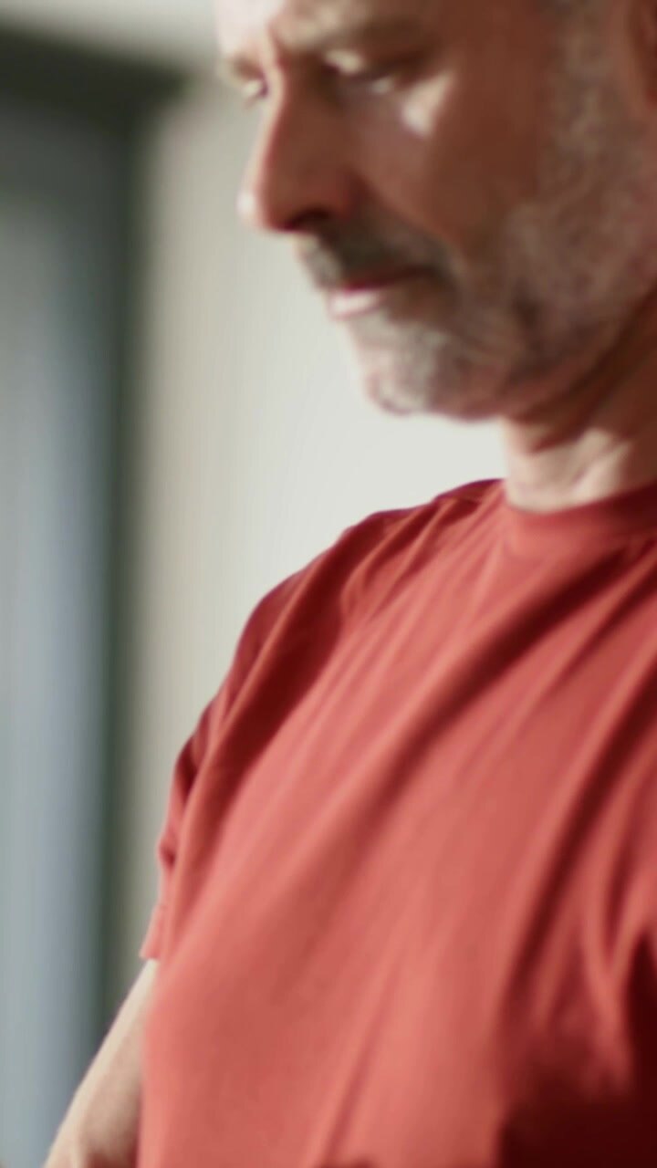 Video verticale: Uomo con camicia rossa che ispeziona, tenendo un vaso alla finestra e inclinando la pianta verso la luce.