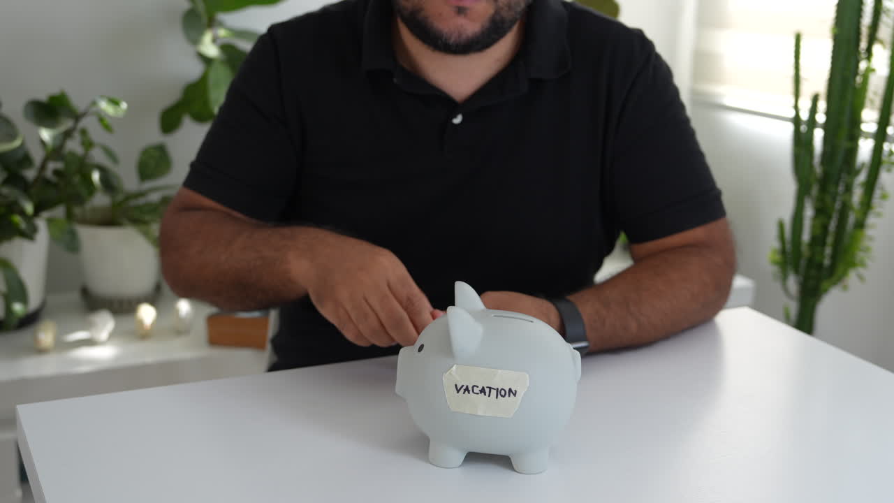 A hispanic man places coins into a piggy bank labeled 'Vacation' on a white table. Sunlight filters through a window, casting soft shadows on plants in the background. Close-up shot