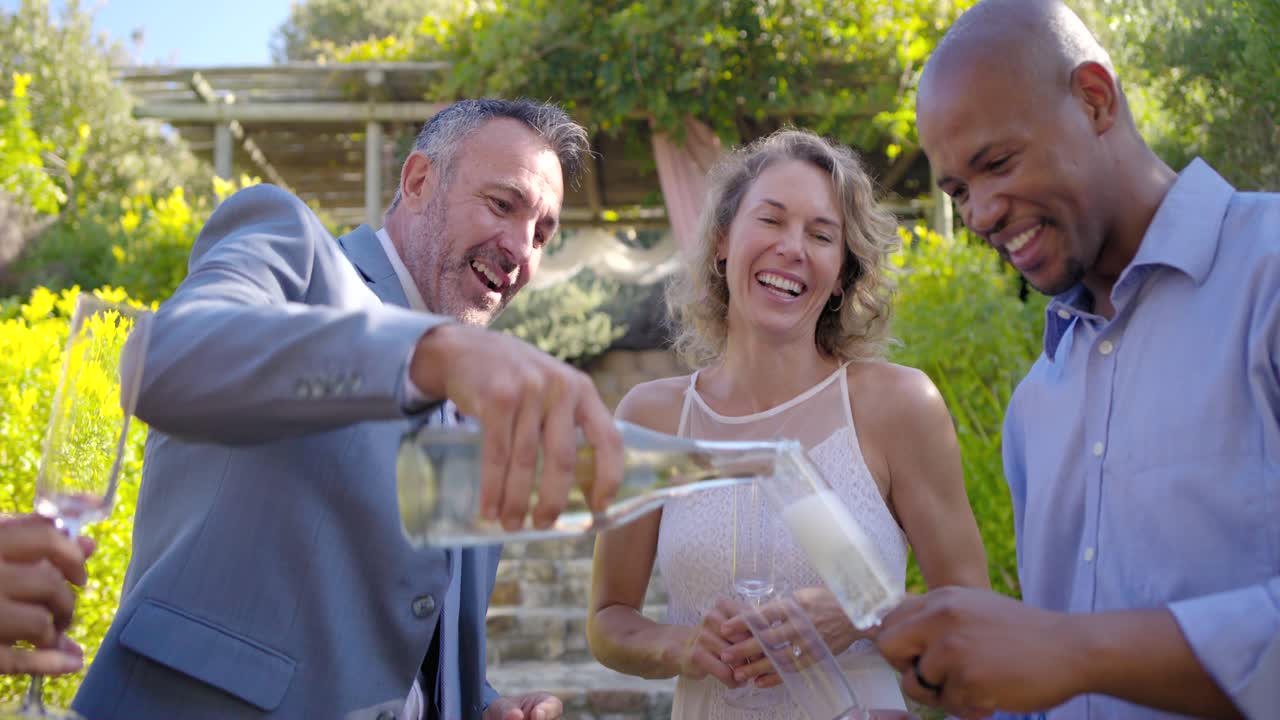 Quattro invitati che ridono, un uomo in abito stappa il tappo, riempie i flûte dalla bottiglia e brinda sotto un pergolato.
