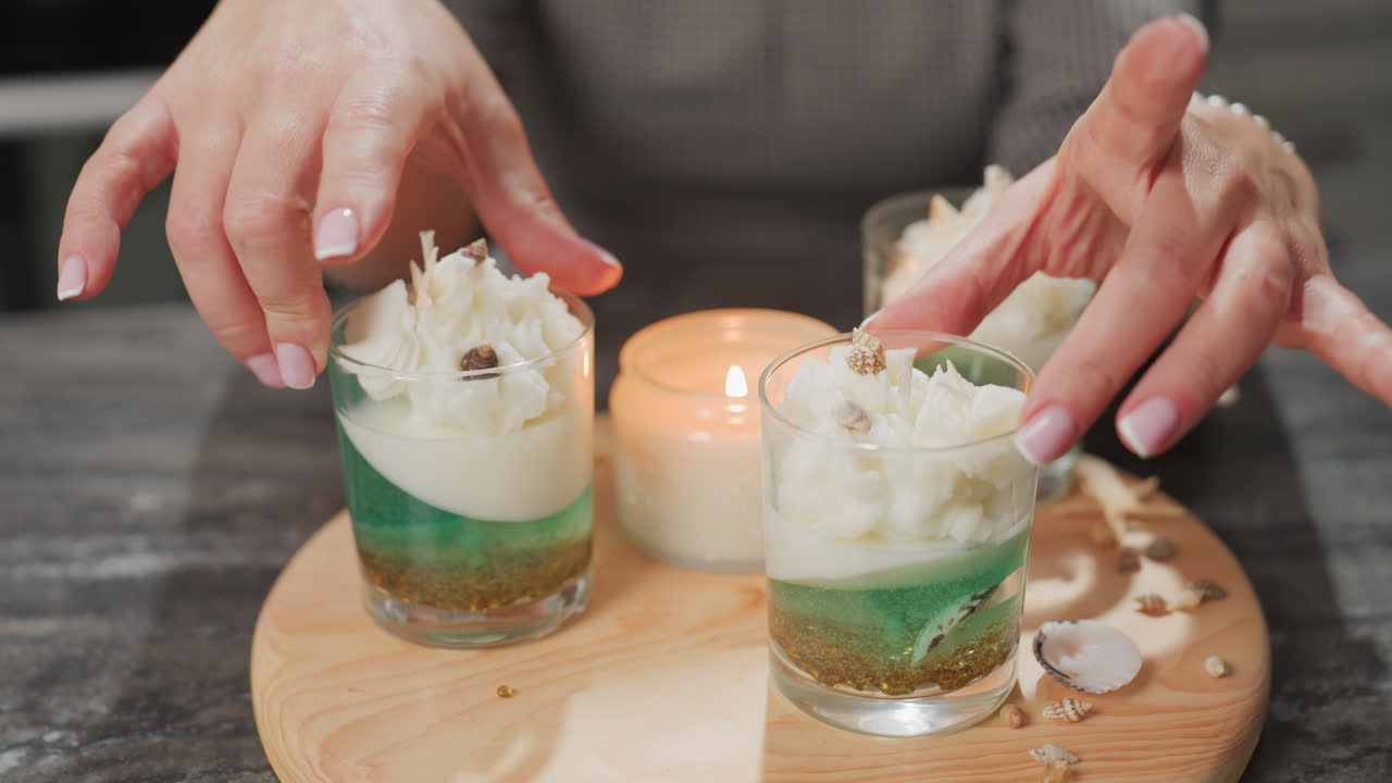 Professional baker with manicured hands gently rotates decorated cupcakes on wooden cake board surrounded by shells and candlelight creating cozy sweet atmosphere filled with creativity passion