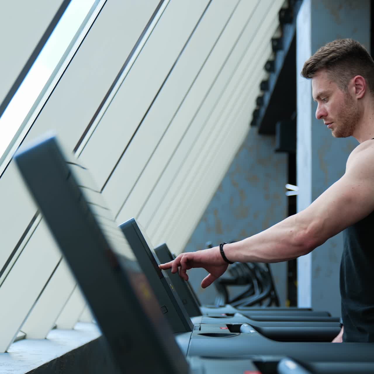 Sportsman in fitness club. Young handsome athlete switches on electronic treadmill in the gym. Muscular man exercising on a simulator. Profile view