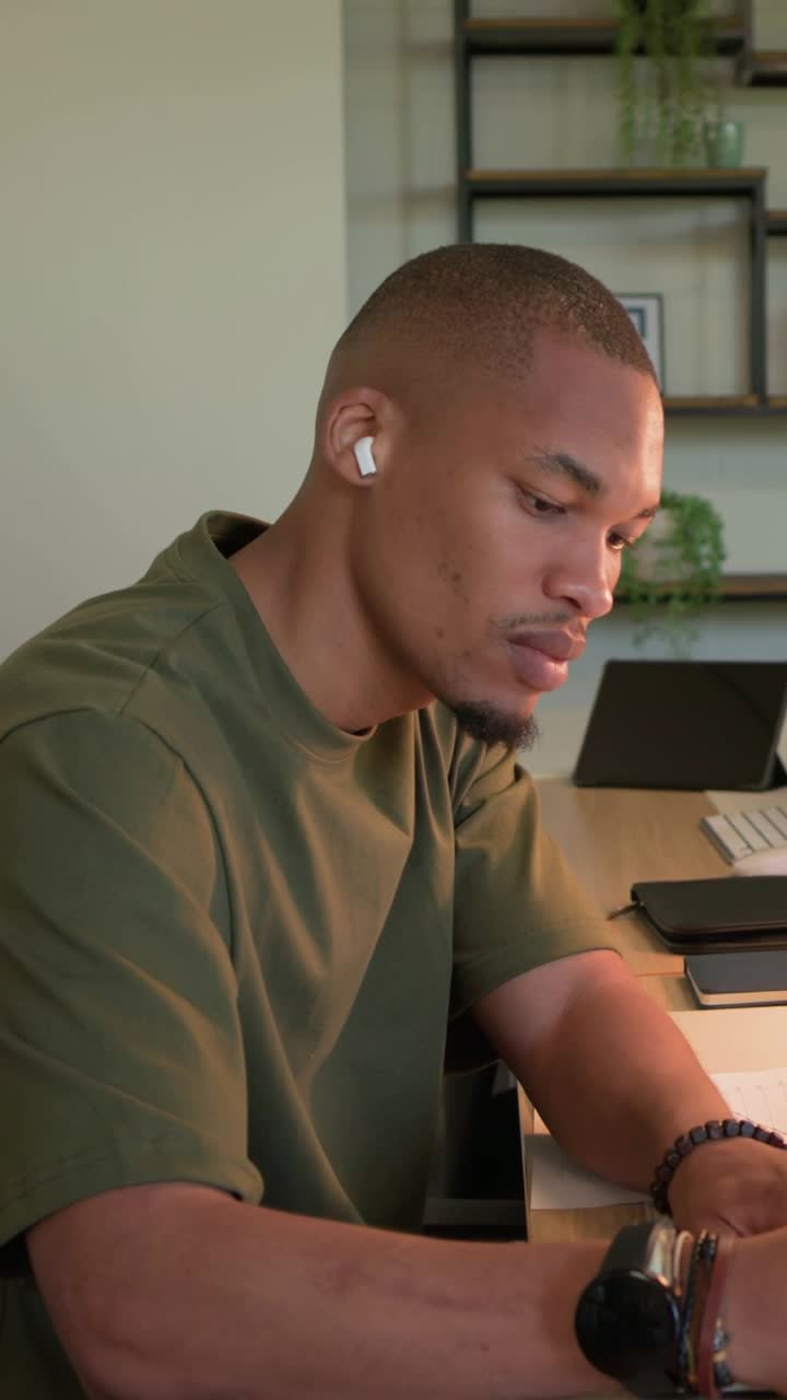 Vertical video: Typing man in olive tee shifting laptop to notebook at desk while camera moving in