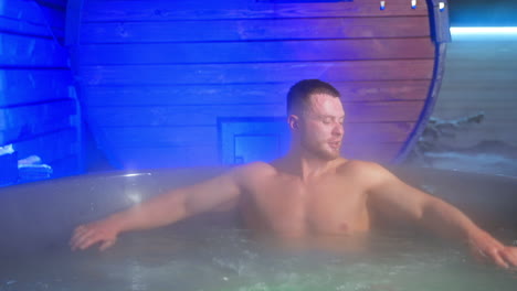 handsome bearded caucasian muscular man sitting in a hot tub outdoors. man is relaxing and washing face with hot water.