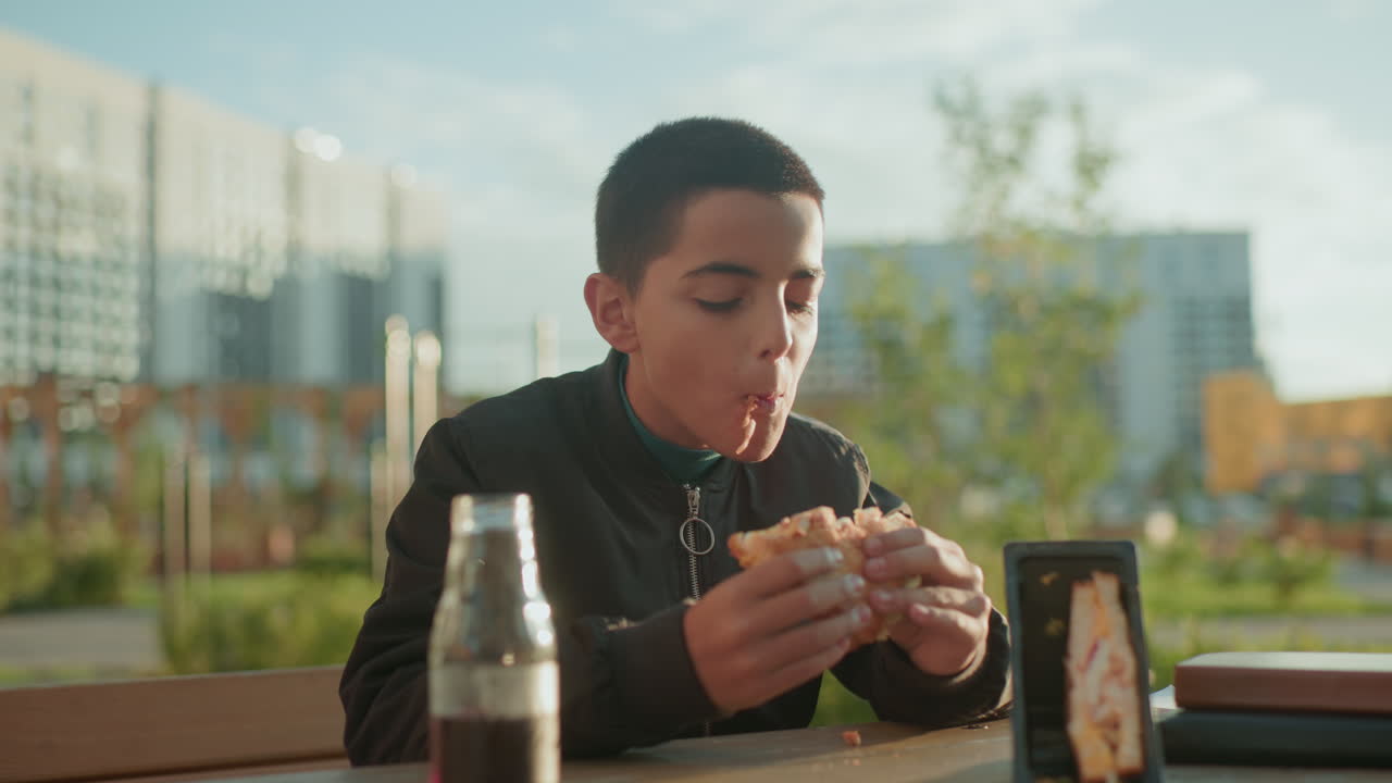 Boy sitting outdoors biting into pizza with juice bottle and sandwich pack on wooden table, enjoying casual snack under soft sunlight with relaxed expression in urban background