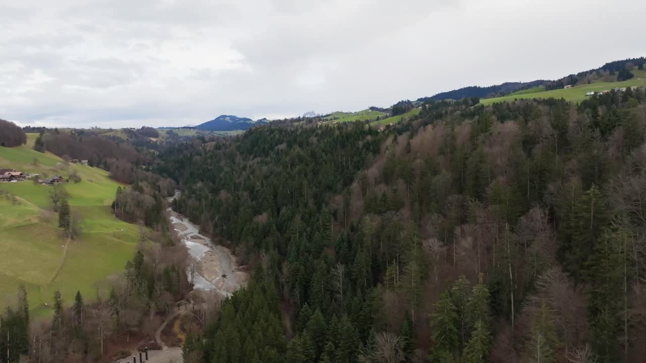 Aerial view of a scenic valley with forest, hills, green fields and small village houses, showing a peaceful rural landscape and natural environment under a cloudy sky
