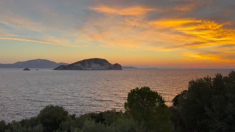 la impresionante puesta de sol sobre la isla de marathonisi en zakynthos, grecia, con nubes vibrantes, reflejos de agua y exuberante vegetación en primer plano.