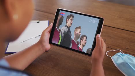 Composite-of-woman-sitting-at-table-with-face-mask,-watching-sports-event-on-tablet