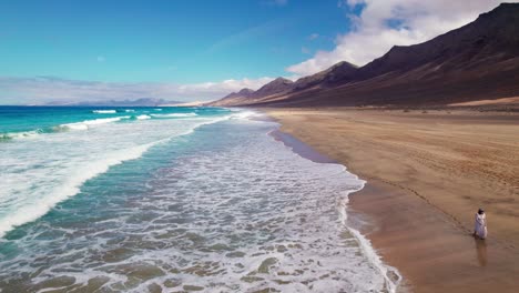 flyover of caucasian woman in white flowing dress as she strolls through the secluded paradise of cofete beach in fuerteventura by aerial drone 4k