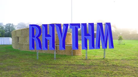 gigantic rhythm text of blue colour on metal fence scaffolding many bales behind the sign and small stage in the background for djs designated area grassy field vibe check entering the summer mood