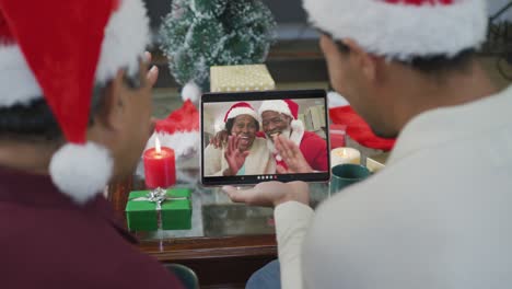 Biracial-father-and-son-waving-and-using-tablet-for-christmas-video-call-with-happy-couple-on-screen