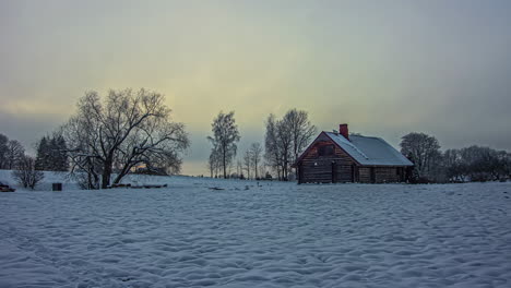 Kleine-Hütte-Im-Schnee-Unter-Orangefarbenen-Wolken-In-Goldenem-Himmel,-Der-Sich-In-Einer-Verschneiten-Landschaft-Mit-Bäumen-In-Einem-Winterzeitraffer-Wolkig-Dreht
