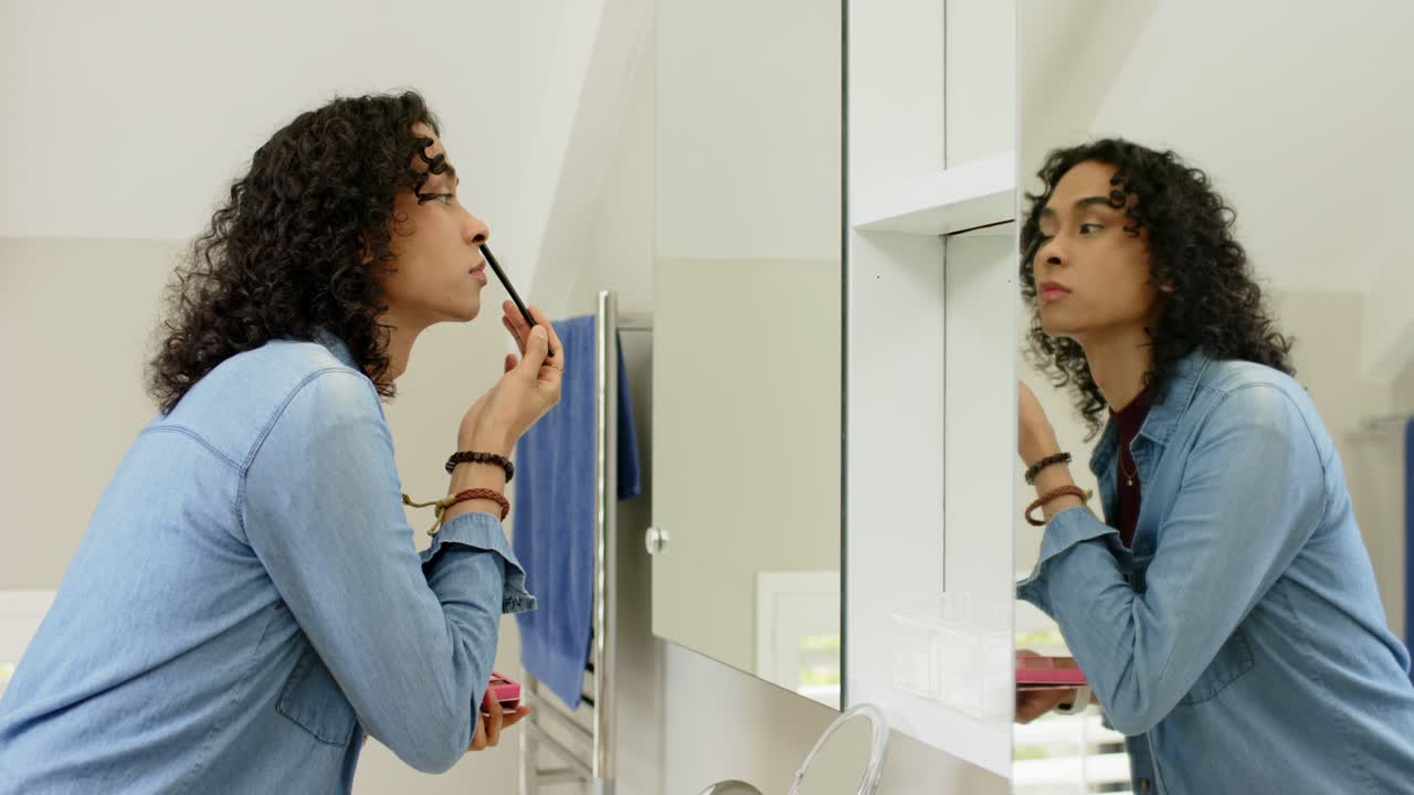 Adulto non binario che entra in bagno, usando una palette e un pennello per delineare le ciglia nello specchio, spazio per il testo.