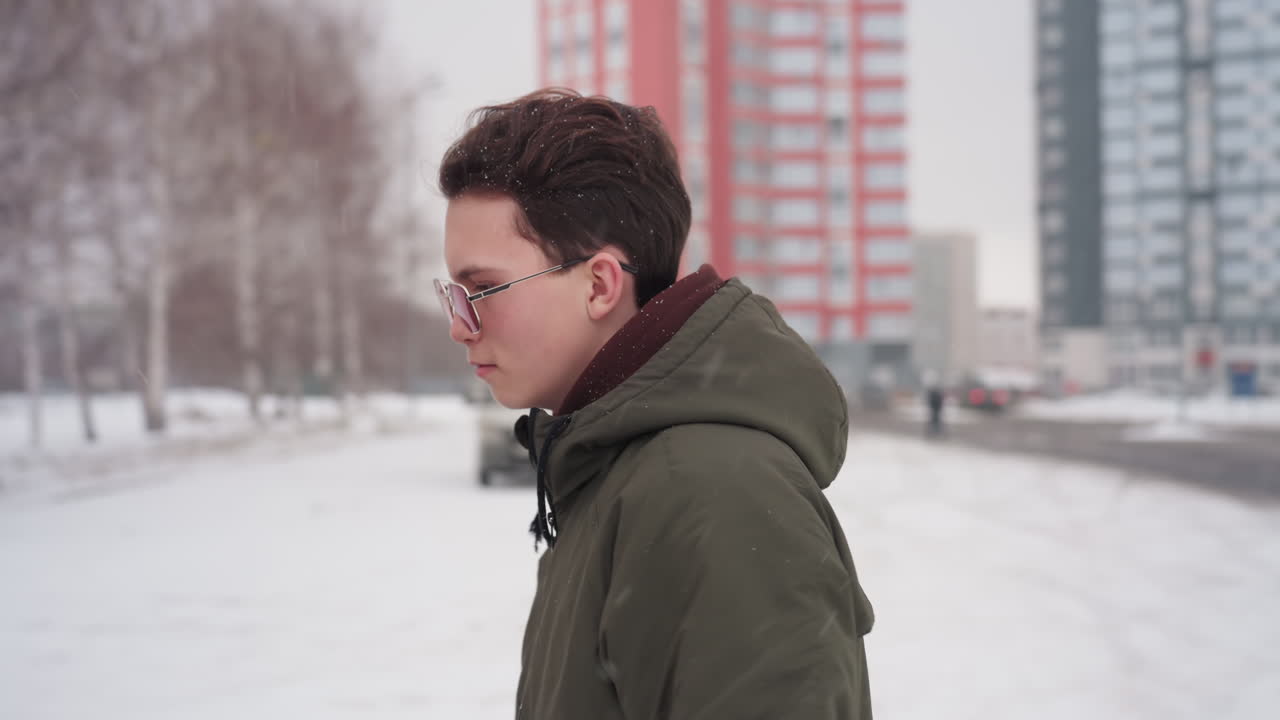 Young man in winter jacket walks through snowy parking lot toward car while snow falls around head distant person visible near tall residential buildings and parked vehicles in cold urban environment