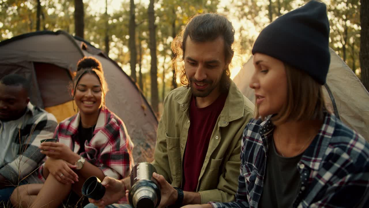 A happy group of four hiking participants are developing and drinking tea from a thermos against the backdrop of tents in a sunny green summer forest. An international group of hikers in a sunny summer forest