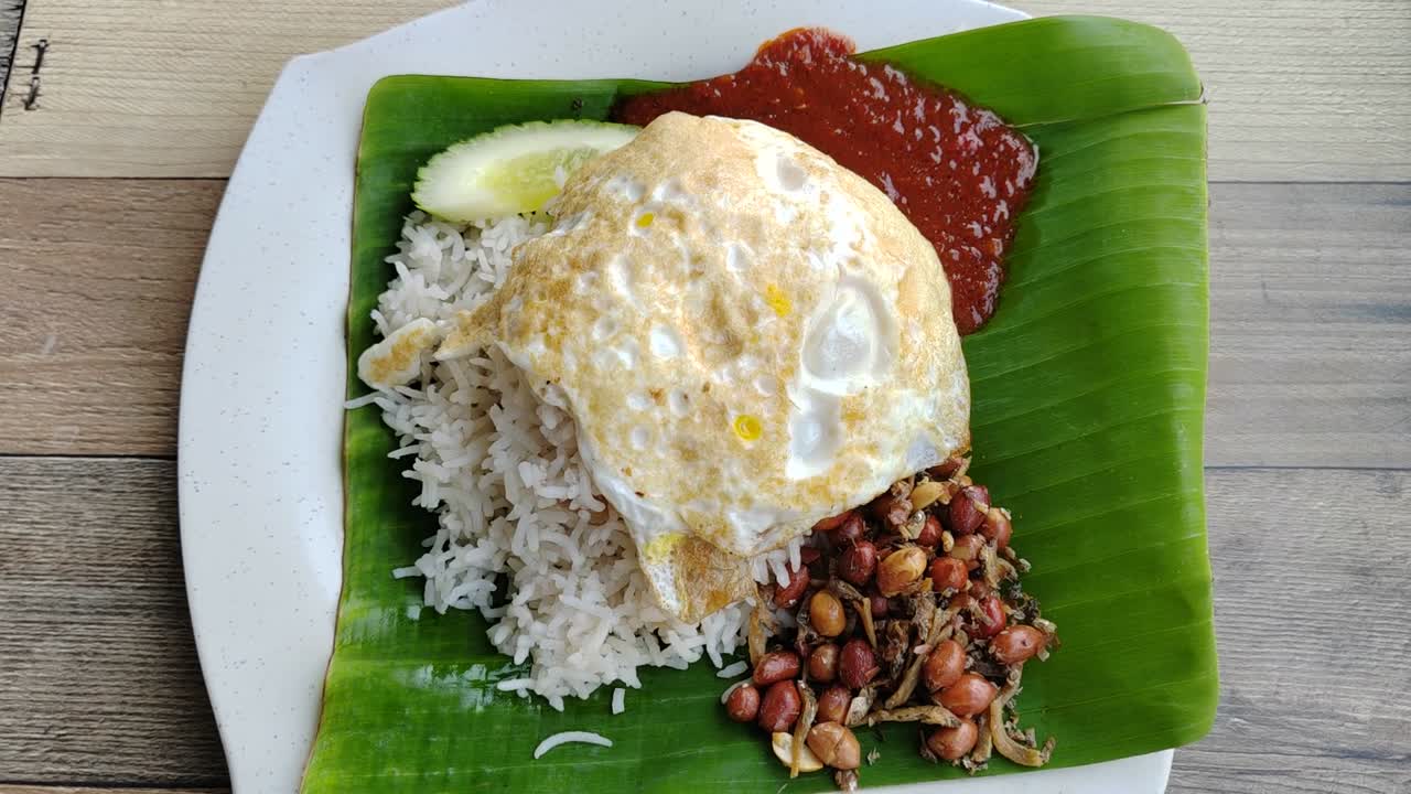 Malaysia's national dish, Nasi Lemak, is plated for serving. The fragrant rice, cooked in coconut milk, rests on a banana leaf. It features classic sides fiery sambal, egg, and crunchy peanuts