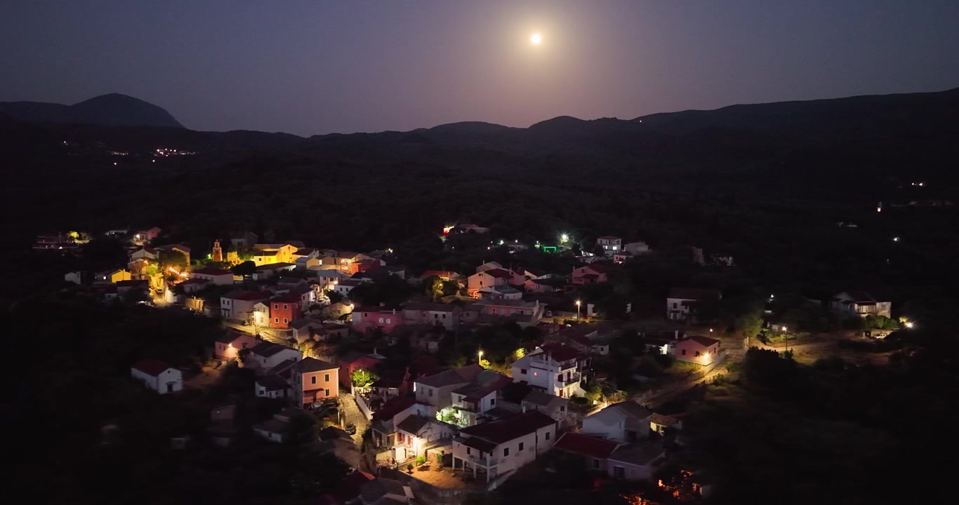Nächtliche Luftaufnahme des Dorfes Agioi Douloi auf der Insel Korfu, Griechenland, mit leuchtenden Häusern unter einem hellen Vollmond.