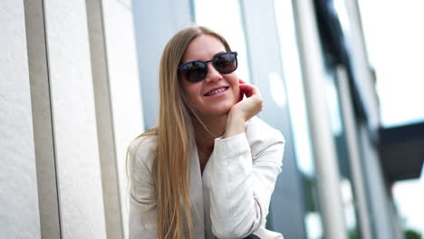relaxed young happy woman sitting outdoors. blonde lady wearing white jacket and black sunglasses smiling to the camera. blurred backdrop.