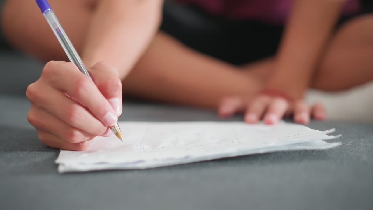 close up of white child sitting cross legged writing on paper with blue pen wearing red rope bracelet on wrist while on sofa during focused learning activity