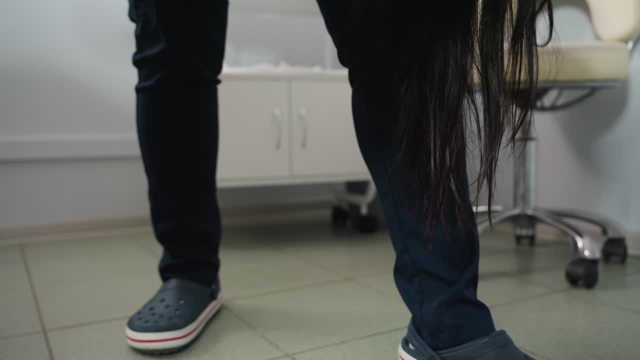 Beauty technician wearing clinical crocs moves beside spa bed as long dark hair strands fall from client onto tiled floor in bright treatment room with storage and basket under soft lighting