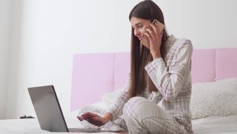 hermosa chica en pijama en la cama hablando por teléfono y trabajando en una laptop, vista lateral del plan general