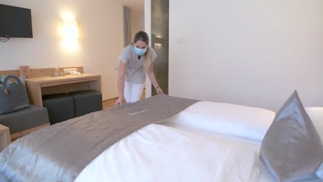 housekeeping with face mask prepares the bed in a hotel room