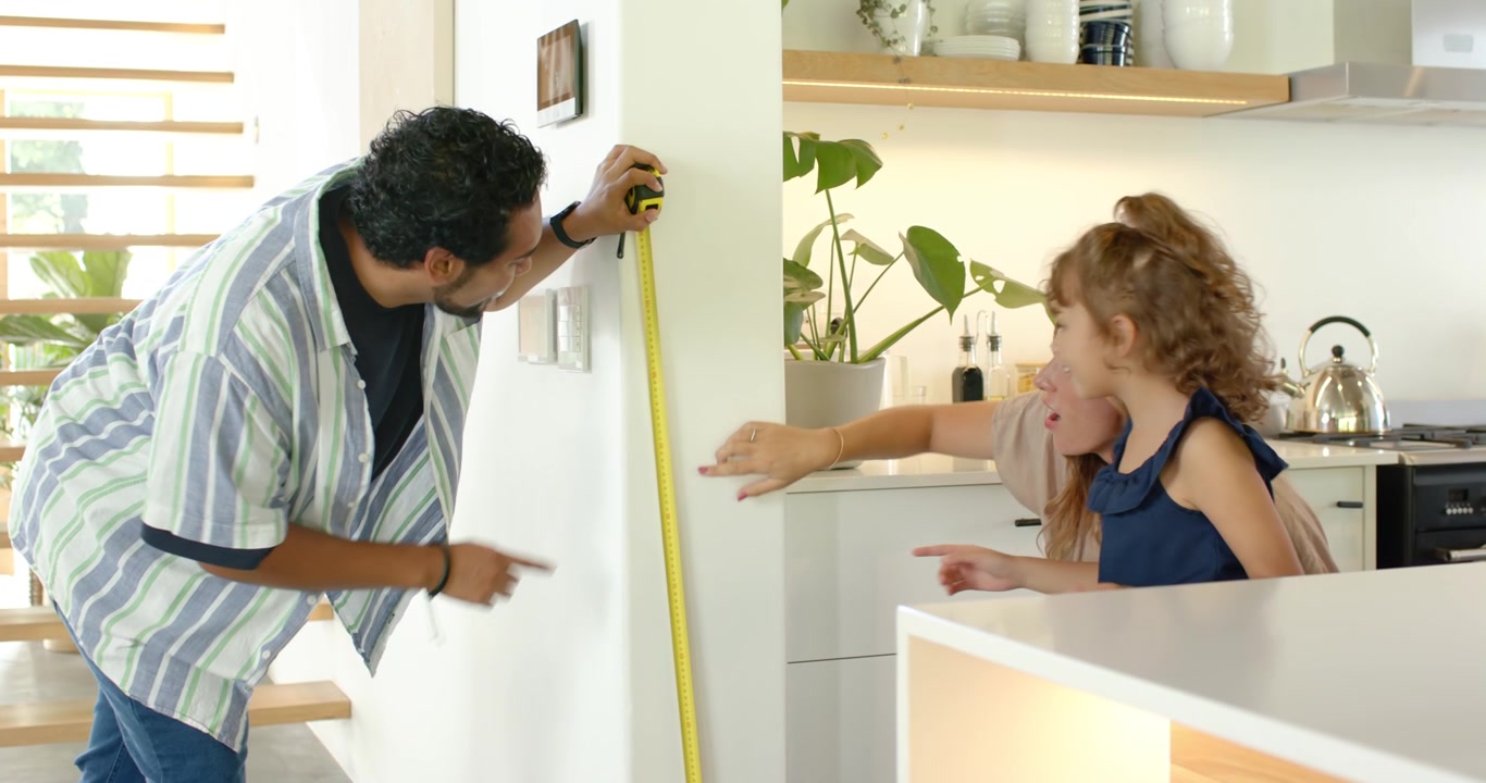 Parents and child measuring height father leaning with yellow tape on kitchen wall marking growth