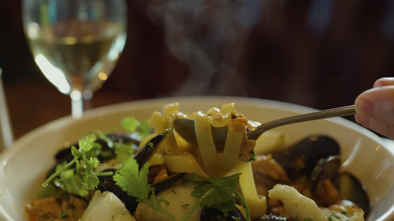 A hand scoops steaming seafood chowder with a spoon beside a glass of white wine in warm, natural light. Macro close-up, shallow depth of field