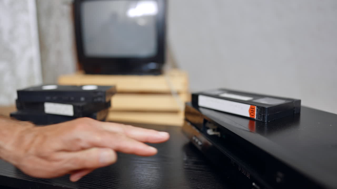 Placing VHS tape into recorder. A hand moves a videotape toward an old VCR with stacked cassettes in the background