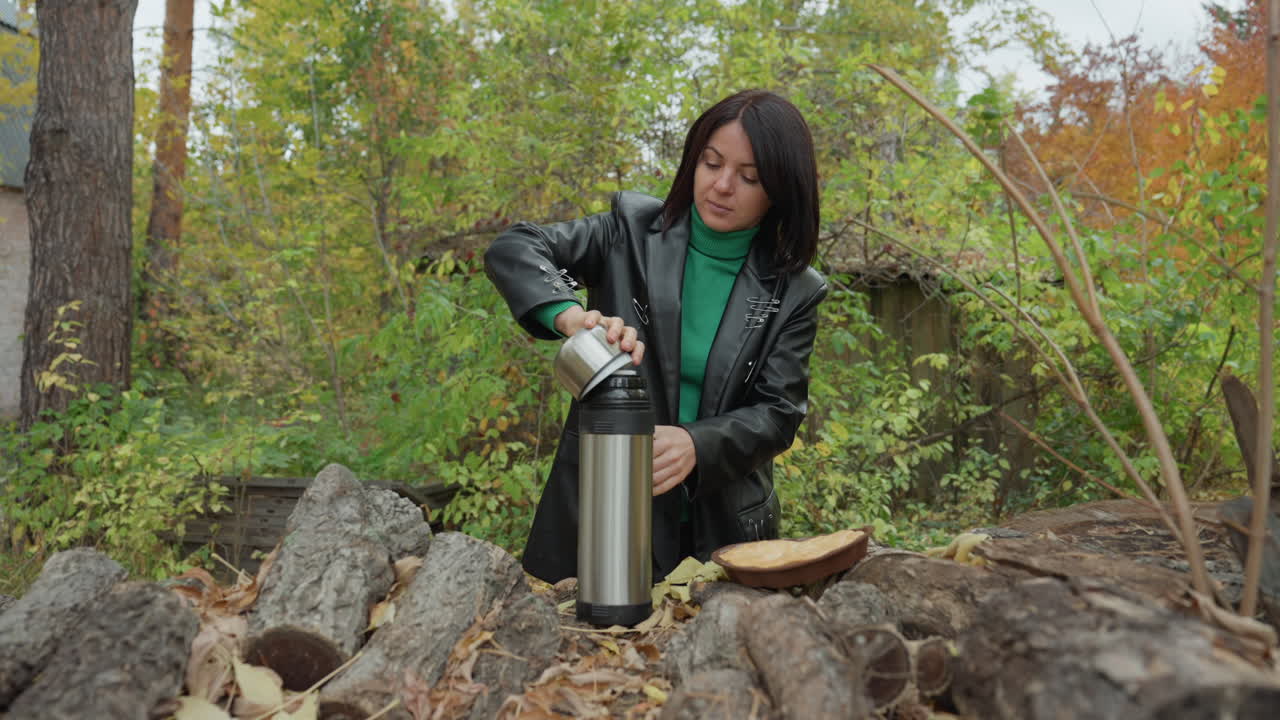woman in leather jacket pouring coffee outdoors, rock musician taking thermos break by log pile, green sweater visible, stainless steel flask and cup on bark, autumn leaves and pine trees
