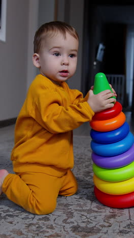 lovely one year old toddler stands on his knees holding a big toy pyramid. boy in orange sport suit drops the toy and crawls up to camera curiously. vertical video