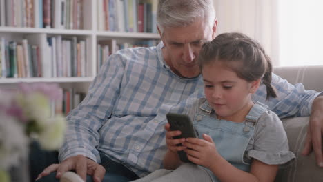 Abuelo-Mostrando-A-Una-Linda-Niña-Cómo-Usar-Un-Teléfono-Inteligente-Enseñando-A-Una-Nieta-Curiosa-Tecnología-Moderna-Niño-Inteligente-Aprendiendo-Teléfono-Móvil-Sentado-Con-El-Abuelo-En-El-Sofá