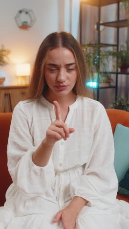 portrait of young woman on sofa presses index finger to lips makes silence hush gesture sign at home