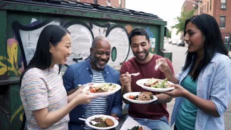 a group of friends enjoying a casual outdoor meal together near a colorfully graffiti-decorated dumpster, celebrating food and friendship in a vibrant urban setting