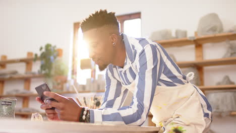 happy african american man with beard using smartphone and smiling in pottery studio, slow motion