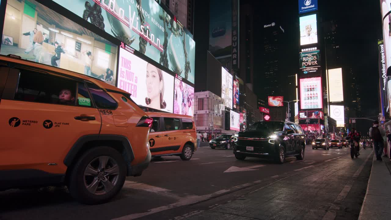 Yellow taxi drives through Times Square at night with glowing billboards and city traffic around