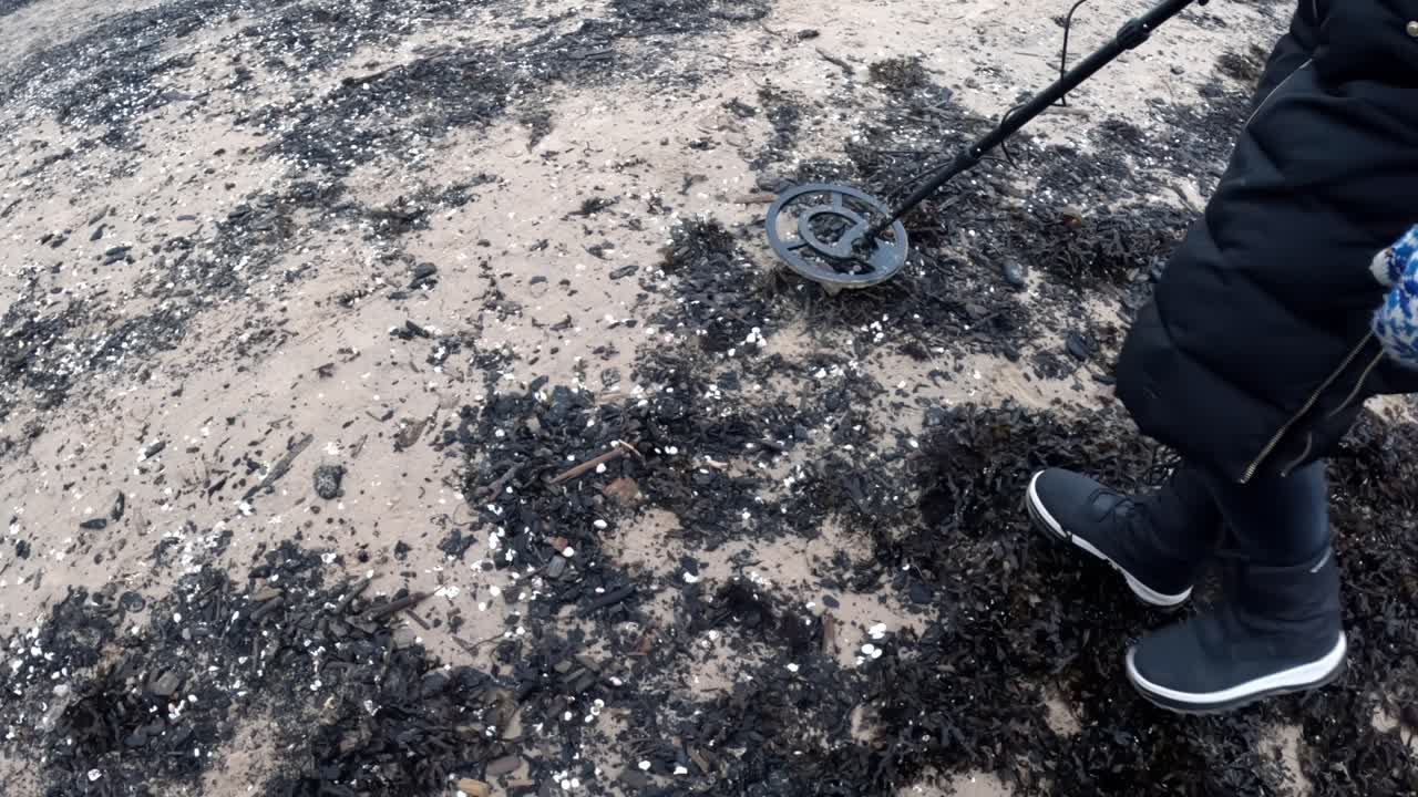 Primo piano di una persona che passa un metal detector su chiazze di alghe scure su una spiaggia fredda, con stivali invernali che calpestano con cura la riva coperta di detriti
