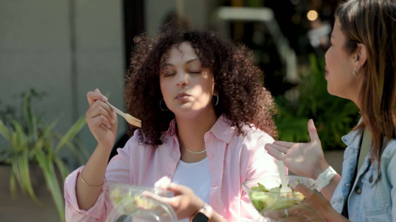 African American women eating at sidewalk table after friend holding salad while passerby crossing