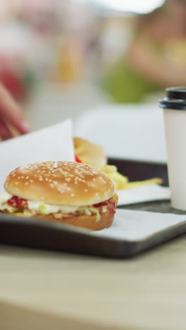 vista inferior del cuerpo de la dama en vestido rosa llevando una bandeja con hamburguesa, papas fritas y bebida camina hacia su mesa, la coloca y la ajusta antes de sentarse, otras personas están en el restaurante