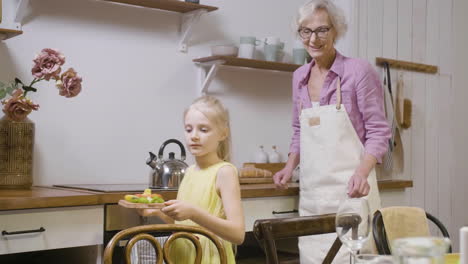 little girl helping her grandmother to set table for dinner and bringing a tray with fresh food