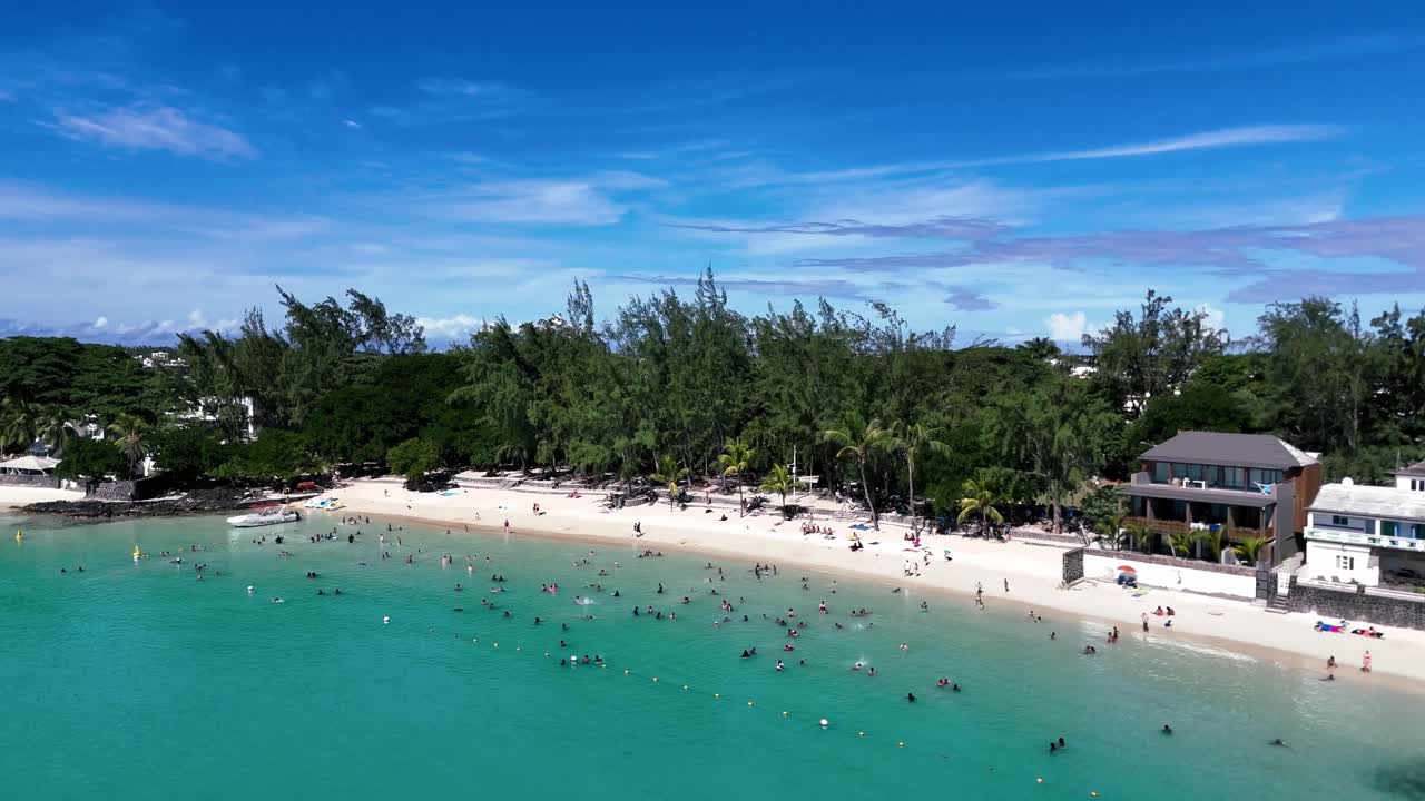 Aerial view of Pereybere Beach, Mauritius from a sweeping side angle, crystal-clear lagoon, lush tropical trees, and lively beach scene under a vibrant blue sky.