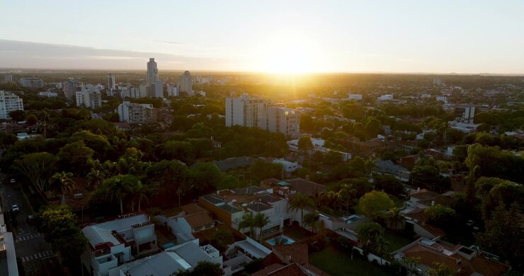 Nascer do sol sobre Assunção, Paraguai, com luz solar quente iluminando o horizonte e bairros residenciais. Atmosfera matinal calma com vegetação, edifícios modernos e paisagem urbana em crescimento