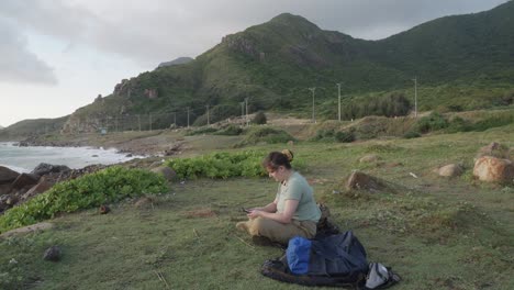 Mujer-Sentada-En-La-Orilla-Del-Mar-Leyendo-Un-Libro-En-Un-Dispositivo-Kindle-En-La-Isla-Con-Dao,-Vietnam