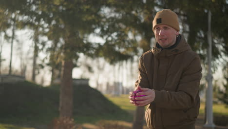 a man dressed in a brown jacket is playing outdoors near trees, catching a purple rotating toy as it floats in the air