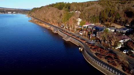 Sunrise-glows-on-a-quiet-lakeside-road-with-nearby-houses,-aerial-side-view