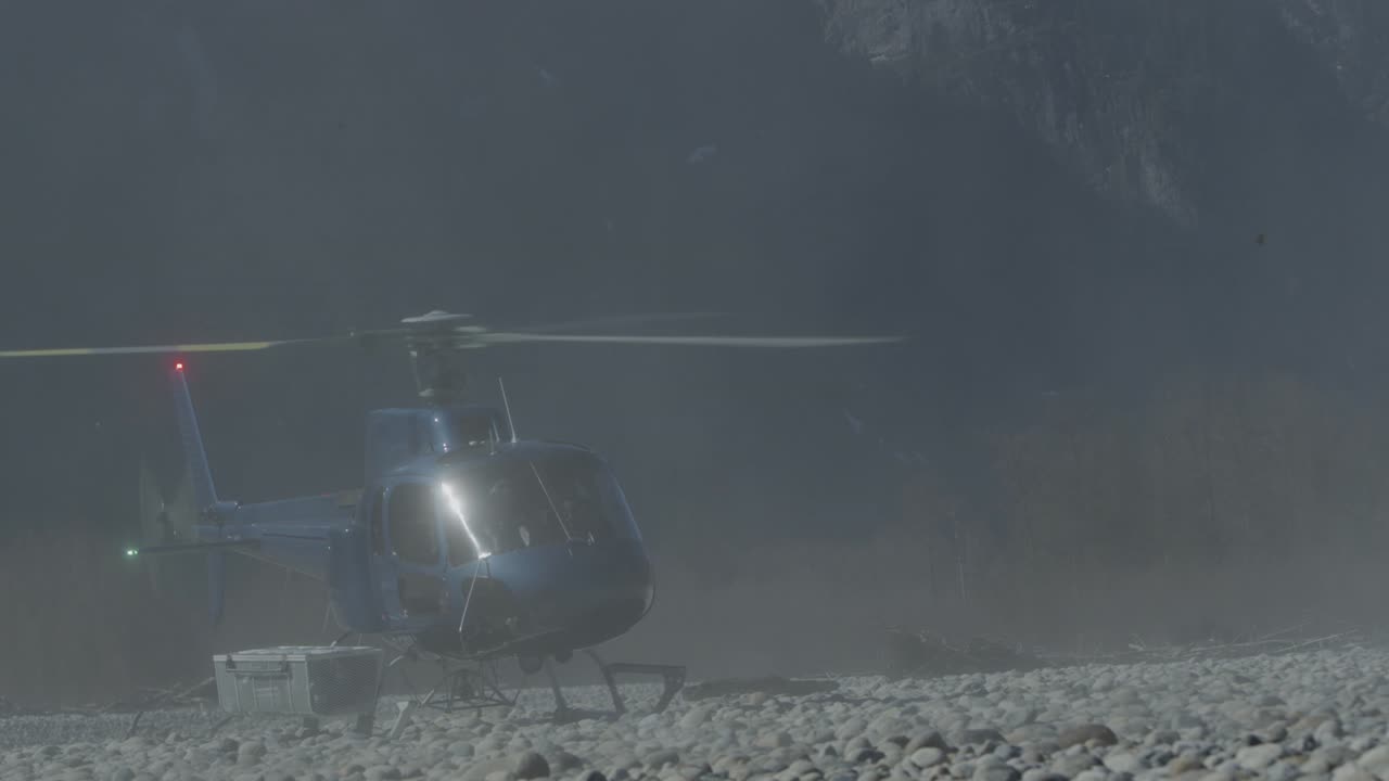 Blue Helicopter landing on rocky beach on river with mountain in background in slow motion 4K Footage