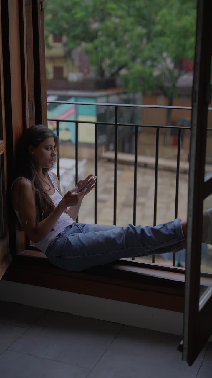 una mujer joven relajándose en un asiento de la ventana, usando un teléfono inteligente en un día lluvioso.
