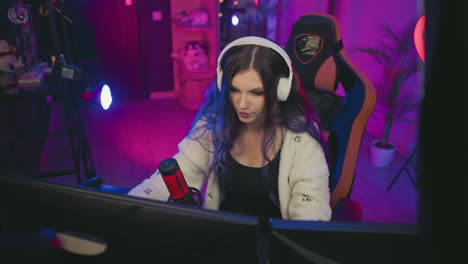 lady wearing white headphones seated on gaming chair in vibrant neon-lit studio playing video game on dual monitors with microphone on desk and pink heart light glowing in background near drum set
